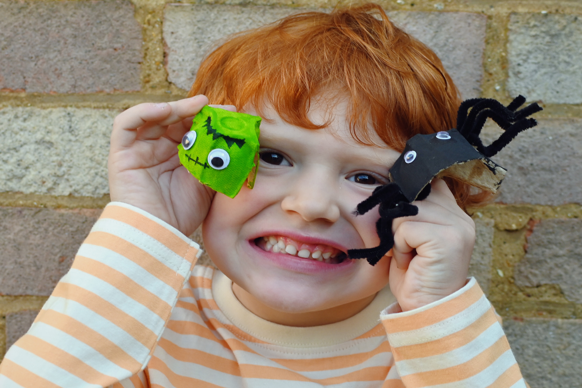 Image shows a child smiling and holding up two Halloween egg carton craft creations.